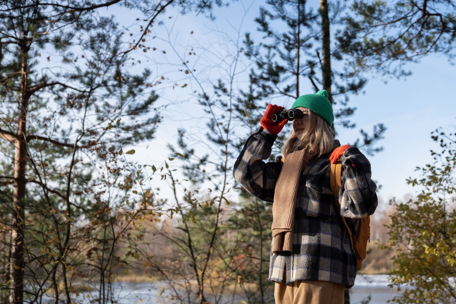 A woman looking through a pair of binoculars in a wooded area by water.