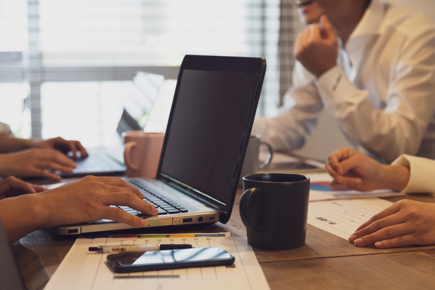 Close-up of people working in an office, with a laptop, paperwork, and mug on the desk.